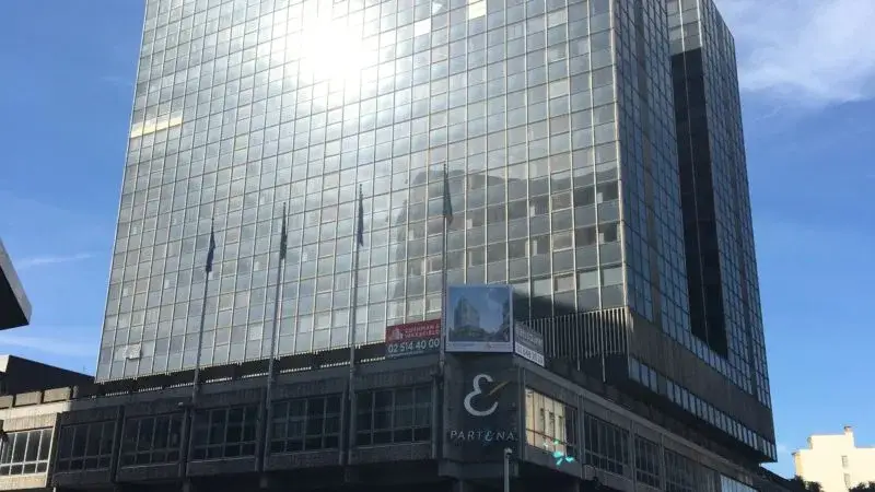 Groot kantoorgebouw met volledig glazen gevel, waarin het zonlicht reflecteert. De onderste bouwlagen bestaan uit een donkerder structuur met zuilen en een hoekbord met bedrijfslogo.
