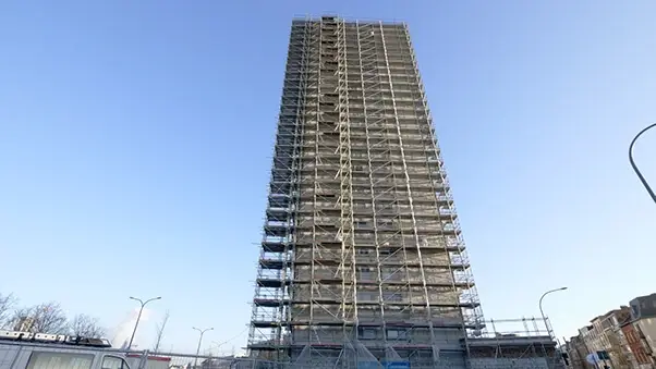 Hoogbouwgebouw volledig ingepakt in een uitgebreid steigersysteem, gefotografeerd vanop straatniveau. De stellingconstructie omhult de volledige gevel.