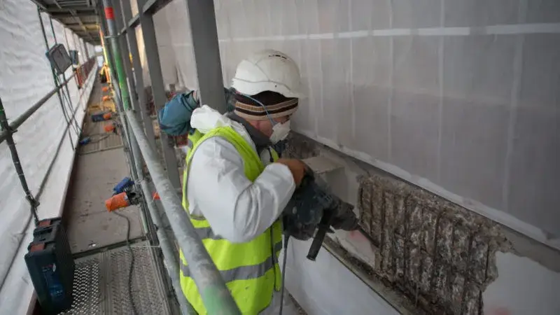 Arbeider op een stelling voert betonherstel uit aan een gevel, met veiligheidshelm, masker en fluohesje. De beschadigde betonzone is opengelegd en de wapening is zichtbaar. De werkomgeving is afgeschermd met beschermfolie.