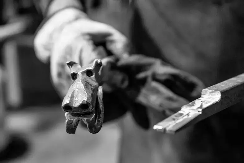 Detail van ambachtelijk smeedwerk waarbij een smid een metalen ornament met dierfiguur vormt, met traditionele smeedgereedschappen in het gespecialiseerd smederij‑atelier van het Renotec ambachtencentrum.