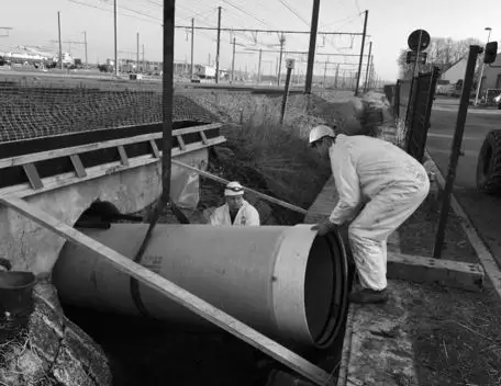 Installatie van een grote betonnen rioolbuis aan een duiker, waarbij twee medewerkers in beschermende werkkledij de leiding positioneren en begeleiden tijdens ondergrondse infrastructuur‑ en rioolwerken langs een spoorweglocatie.