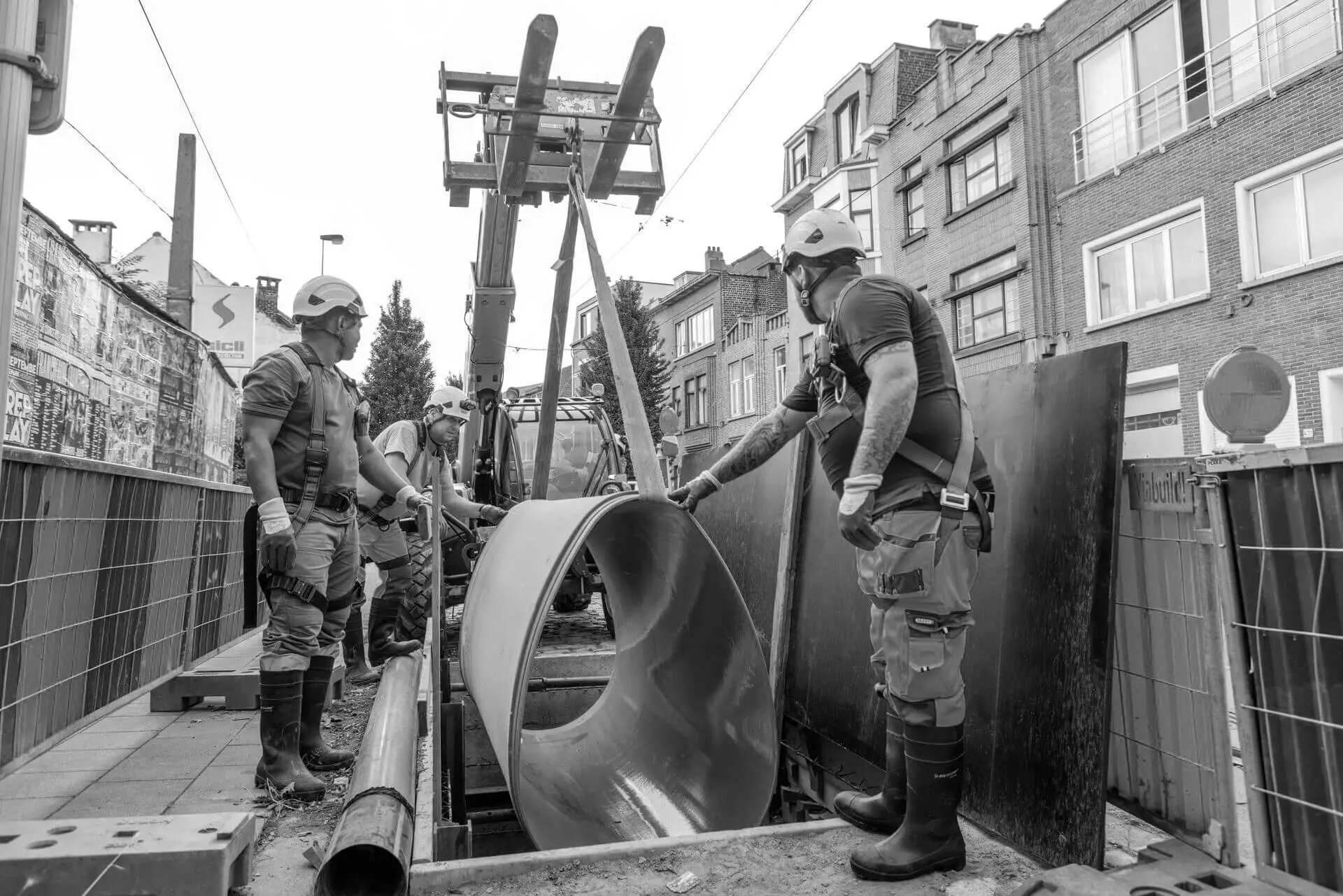 Plaatsing van een groot GVK‑schaaldeel in een open sleuf langs een straat, met een hijsinstallatie op een graafmachine en een team van infrastructuurwerkers die het element begeleiden tijdens een riolerings‑ en ondergrondse renovatie.