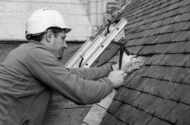 Dakwerker plaatst of herstelt leien op een historisch dak met behulp van een hamer en haak, naast een ladder en metselwerk, als onderdeel van een gespecialiseerd restauratie‑ en dakrenovatieproject.