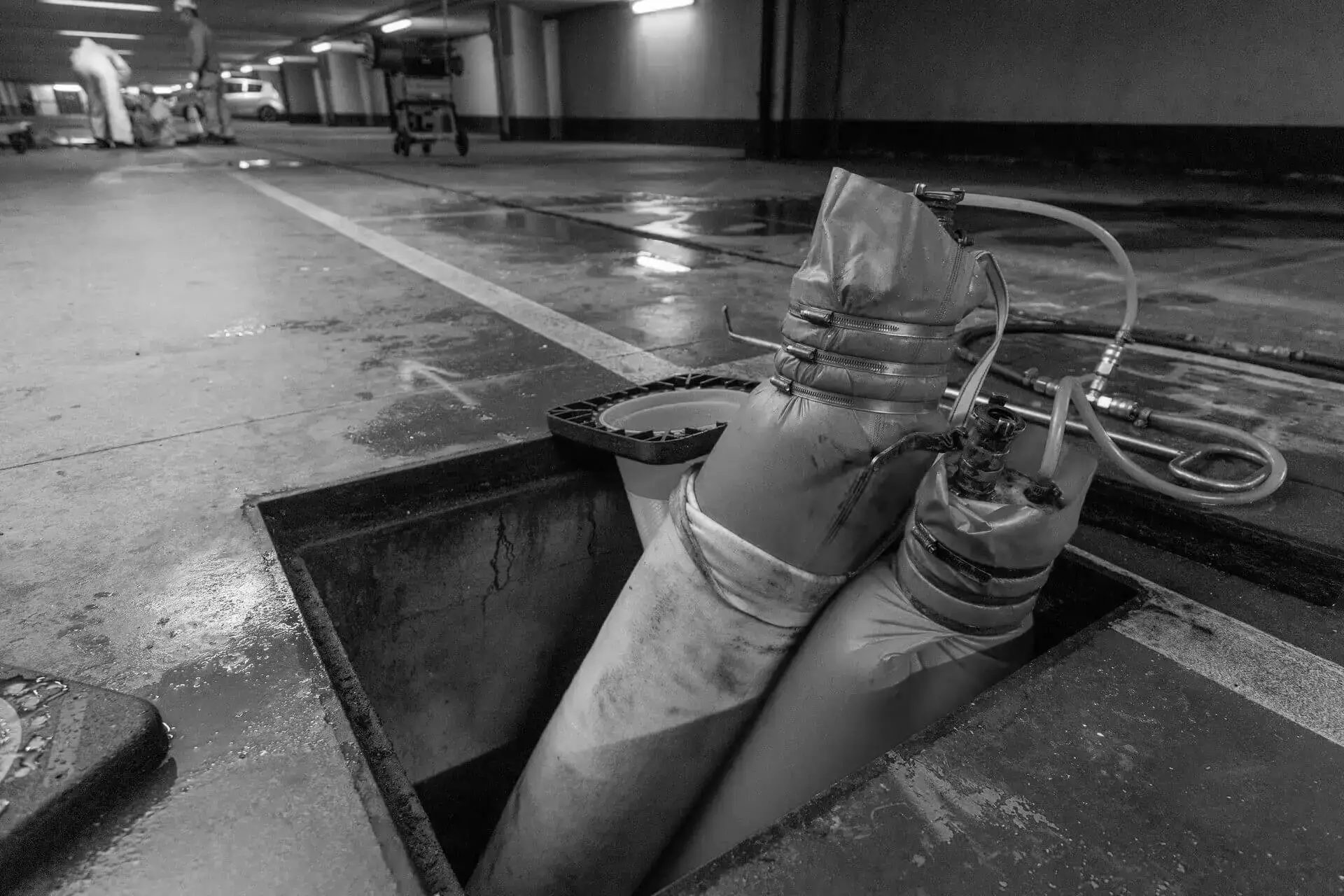 Naaldviltkous die wordt ingebracht via een mangatopening in een ondergrondse leiding in een parkeergarage, met aansluitingen voor lucht- of waterdruk tijdens een kousreliningproces voor rioolrenovatie.
