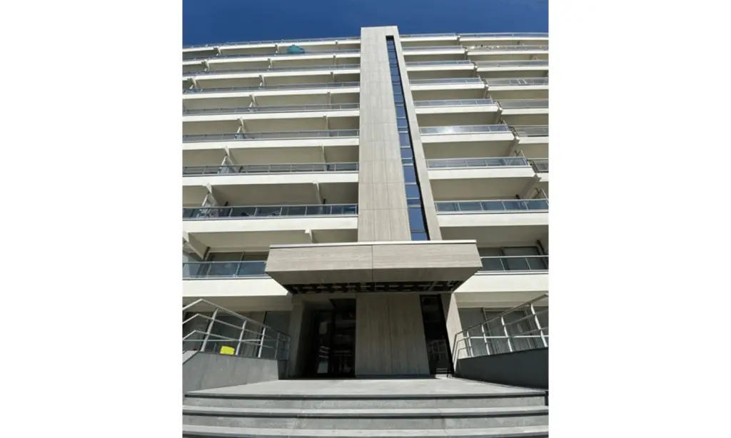 Een modern gebouw met meerdere verdiepingen en glazen balkons, gezien vanaf de onderkant. De ingang heeft brede treden die leiden naar een hoog verticaal architectonisch element boven de deuropening.