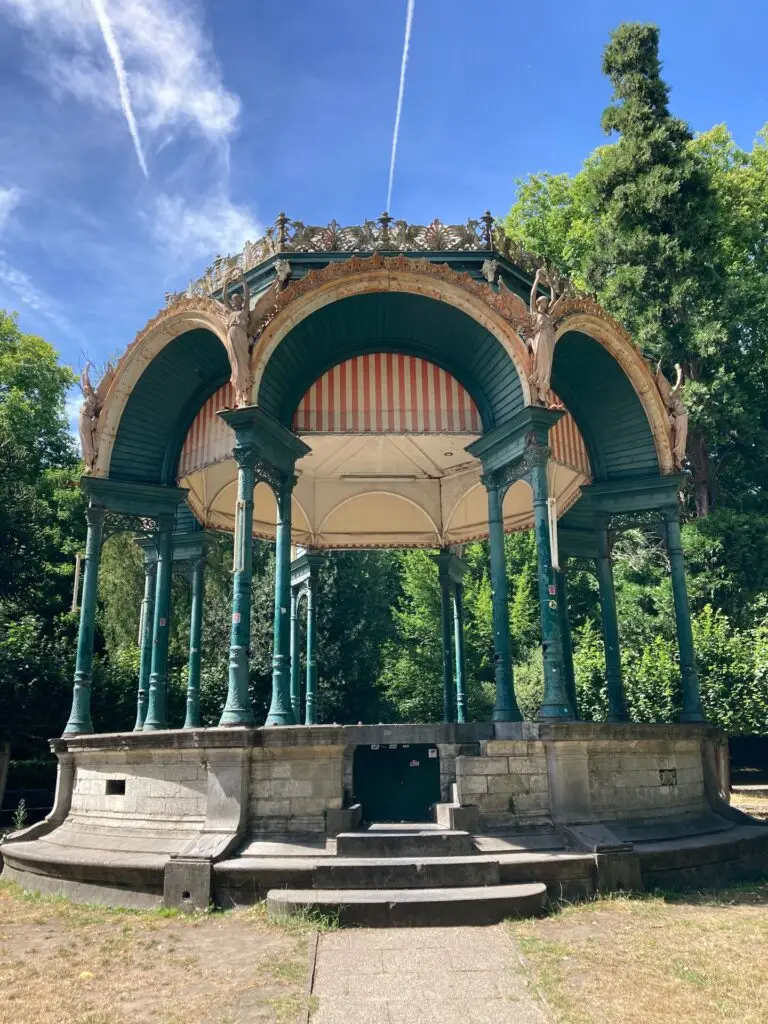 Historische gietijzeren muziekkiosk in een groen park in Gent, met sierlijke bogen, slanke kolommen en decoratieve ornamenten.
