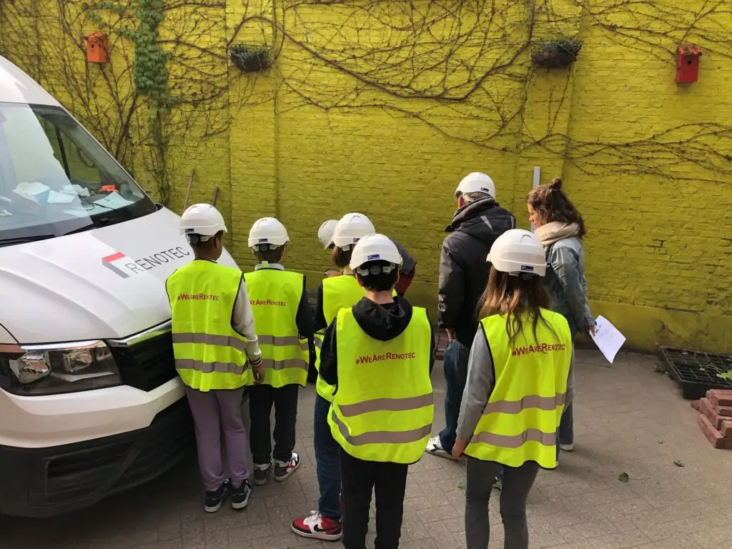 Groep leerlingen met veiligheidshelm en fluohesje krijgt uitleg tijdens beroependag naast Renotec-voertuig, kennismaking met bouw- en renovatiesector op de Merode-school in Berchem